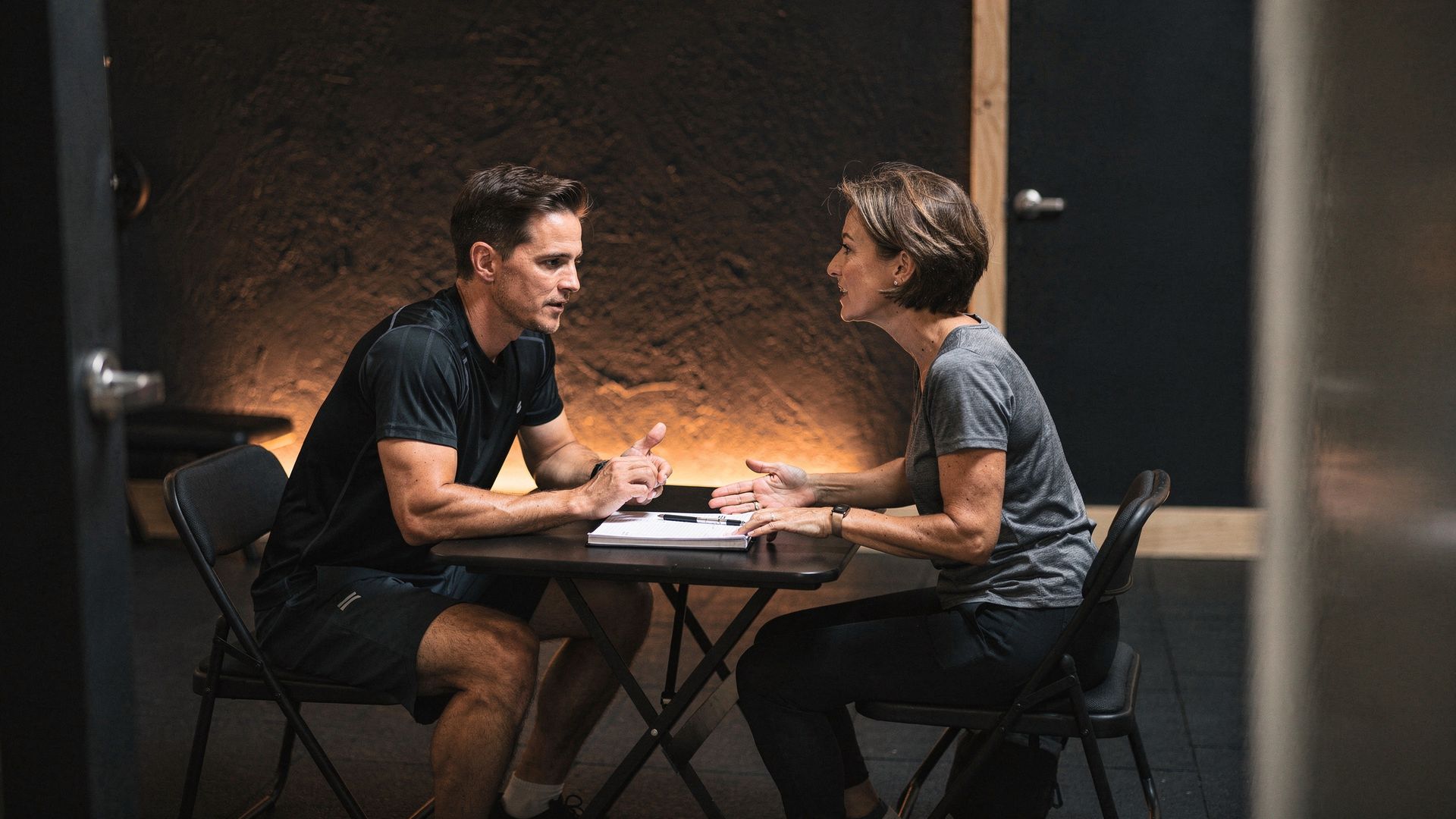 A member and trainer talking in a small dark studio, mid-session